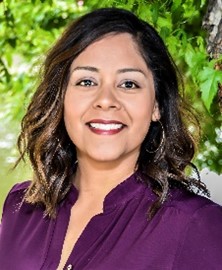 Woman with medium length brown hair, brown eyes and red lipstick smiling in a purple dress shirt in front of a leafy background
